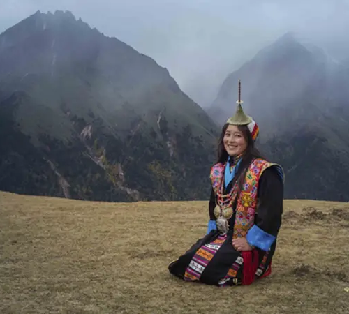Traveler wearing traditional Bhutanese dress posing in scenic Phobjikha Valley with Himalayan mountains in the background.