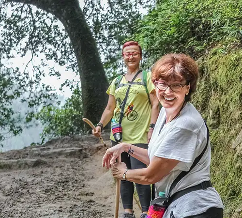 Two smiling female trekkers walking along a lush forest trail with a Bhutan trekking guide, surrounded by moss-covered trees and greenery in Bhutan.