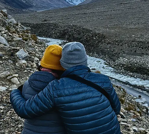 A couple wearing warm beanies and down jackets sits by a mountain stream, enjoying a quiet moment during their Tibet Tour from Australia.