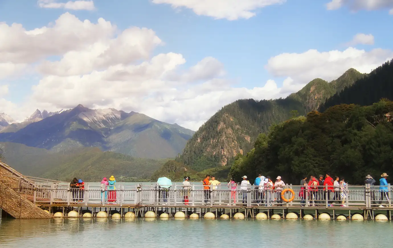 Visitors walk along a lakeside boardwalk at Basomtso National Forest Park in Nyingchi, surrounded by forested mountains during a Tibet adventure tour.