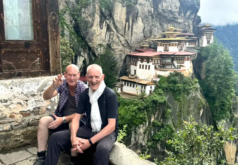 Two travelers sitting on a stone ledge overlooking the iconic Tiger’s Nest Monastery, a highlight of many Bhutan Tour Packages.