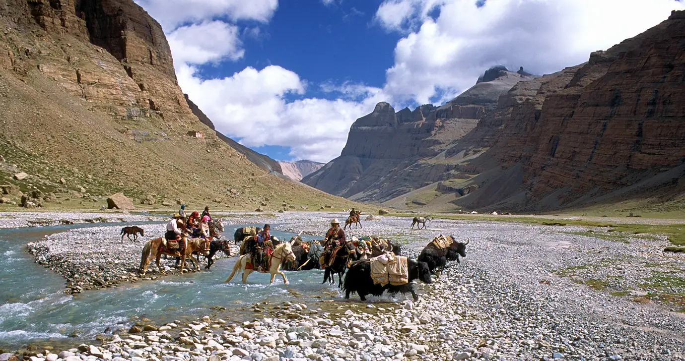 Travelers on horseback and yaks cross a shallow river in a remote Himalayan valley during a Tibet adventure tour near Mount Kailash and the Changthang plateau.