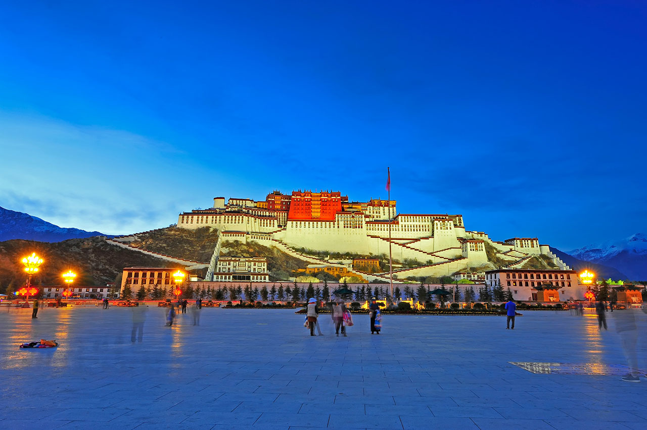 Evening view of Potala Palace illuminated above Lhasa city with visitors walking across Potala Square in Tibet.