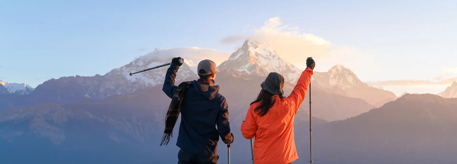 A young couple of travelers celebrating at the Poon Hill viewpoint in Ghorepani, raising their trekking poles toward the Annapurna mountain range at sunrise.