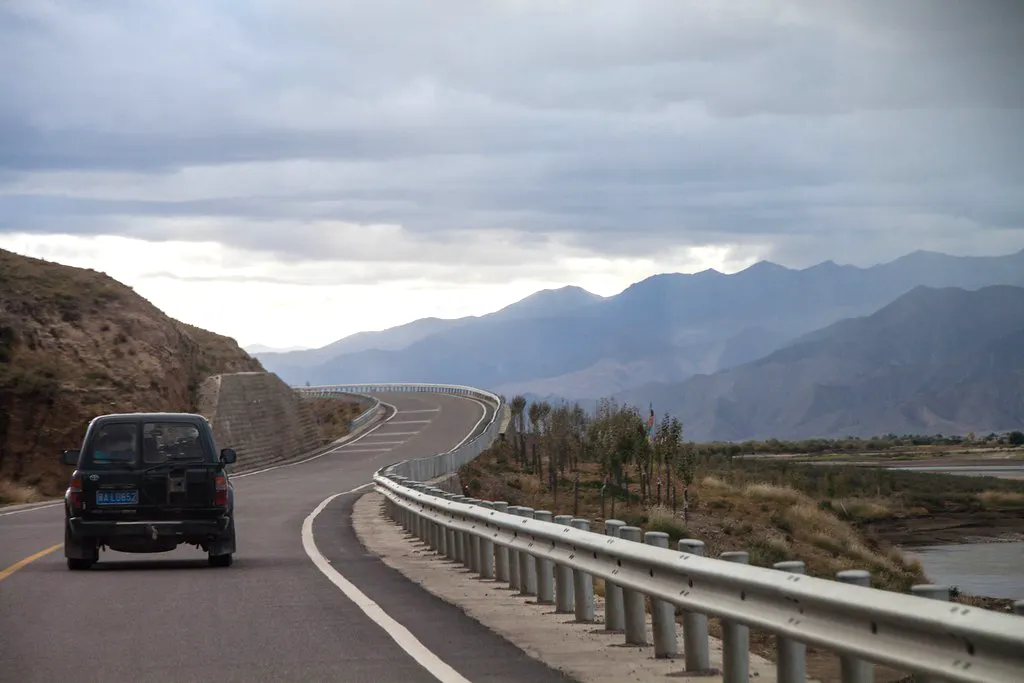 A curved mountain road with guardrails and a vehicle driving during a Road Trip from Lhasa to Shigatse under cloudy skies.
