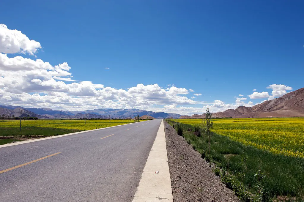 Wide open highway beside green fields and distant mountains during a Road Trip from Lhasa to Shigatse with bright skies and clear visibility.