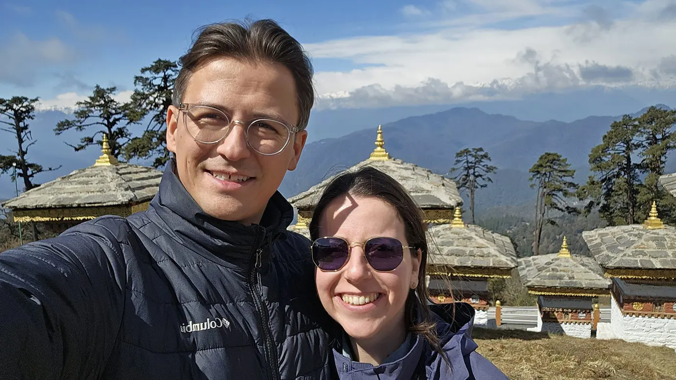A close-up selfie of a couple at Dochula Pass with 108 memorial chortens in the background, a popular scenic stop on Bhutan Tour Packages.