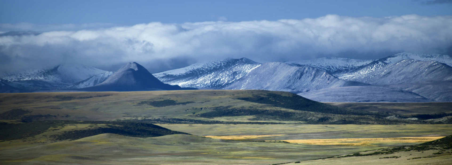 Snow-capped mountain peaks rise above vast, rolling green plains under a thick layer of clouds. This stunning landscape is a centerpiece of a budget Tibet tour.