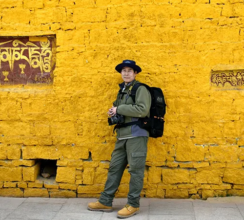 A traveler stands with a camera against a bright yellow wall featuring Tibetan script. He wears layers suitable for the changing Tibet weather by month.