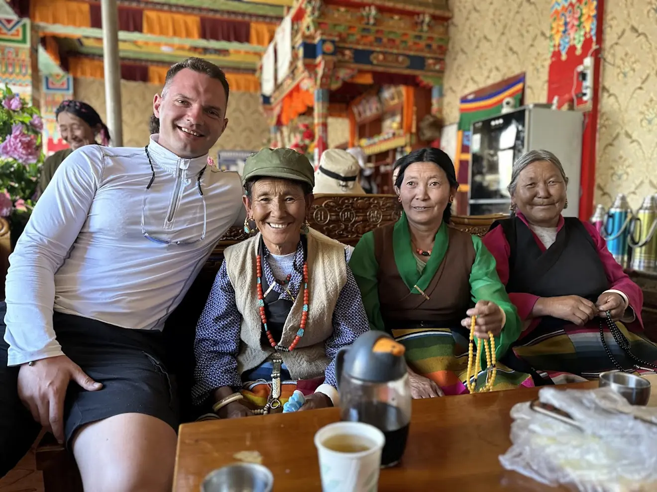 A male traveler sits and smiles with three Tibetan women in a traditional teahouse. This authentic social exchange is a highlight of a Tibet tour from the UK.