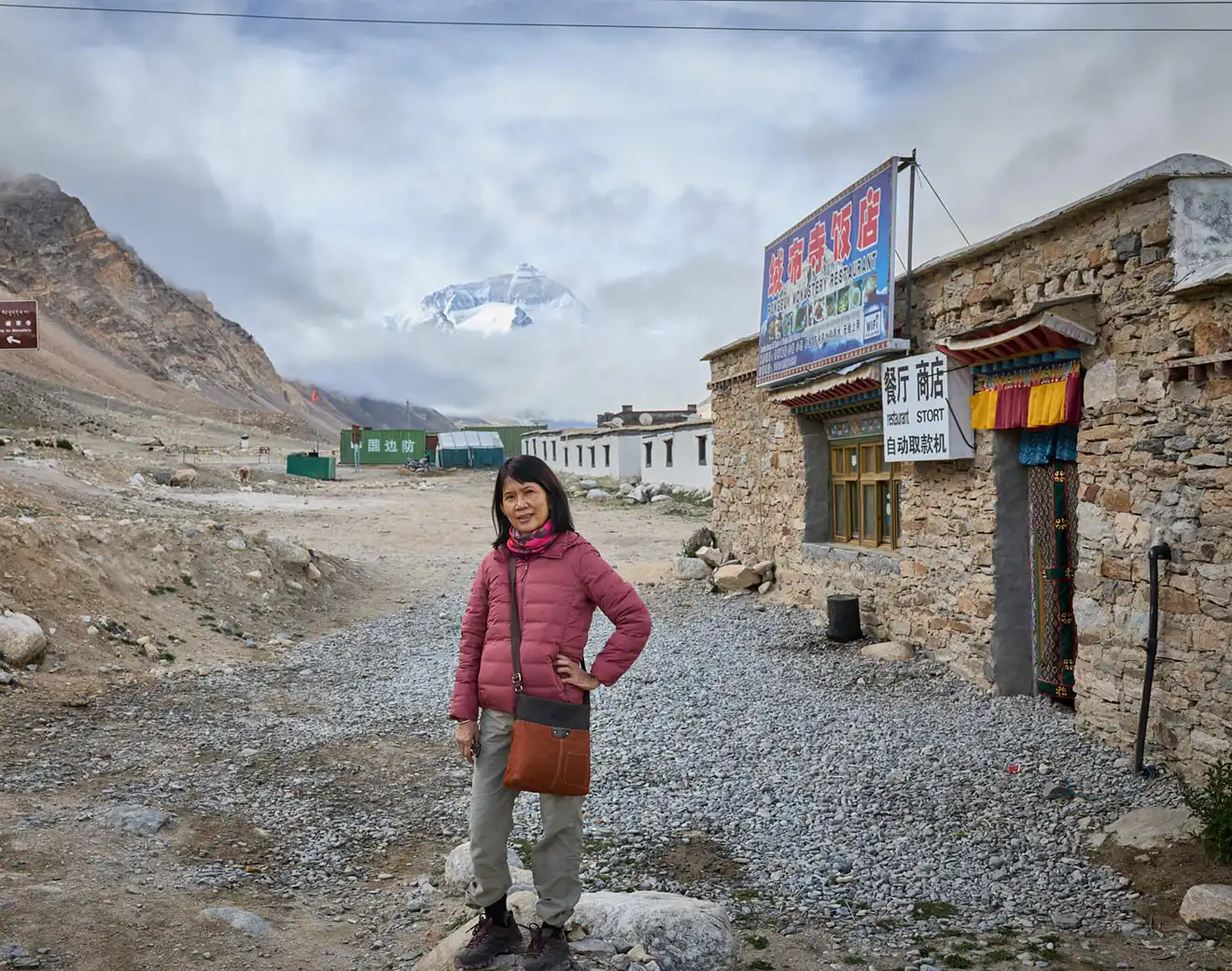 A woman in a pink puffer jacket stands on a rocky path near Rongbuk Monastery with Mount Everest in the background. Seasonal planning is vital for finding the best time to visit Tibet for mountain views.