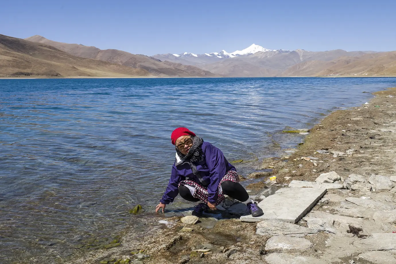 A traveler crouches beside Yamdrok Lake with clear blue water, dry hills, and distant snowy peaks during a scenic stop on a solo Tibet travel route.