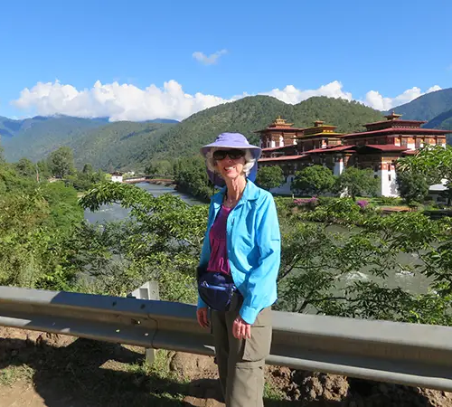 Female traveler standing near Punakha Dzong with river and mountain scenery in the background, showcasing a scenic visit to a historic Bhutan Dzong in Punakha Valley.