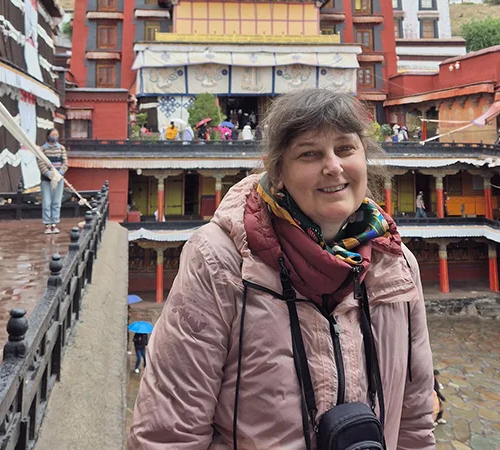 A woman in a pink jacket smiles at a red and yellow monastery courtyard, showcasing a personal experience on a Tibet Buddhist Pilgrimage Tour.