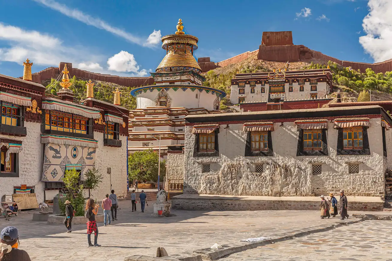 Travelers walk through a historic Tibetan square on a Tibet Tour with Yamdrok Lake and Gyantse, surrounded by traditional buildings, a large stupa, and hillside fort walls.
