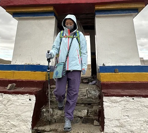 A hiker in a hooded jacket and purple trousers descends stone steps from a traditional stupa. Choosing the best time to visit Tibet ensures comfortable weather for trekking.