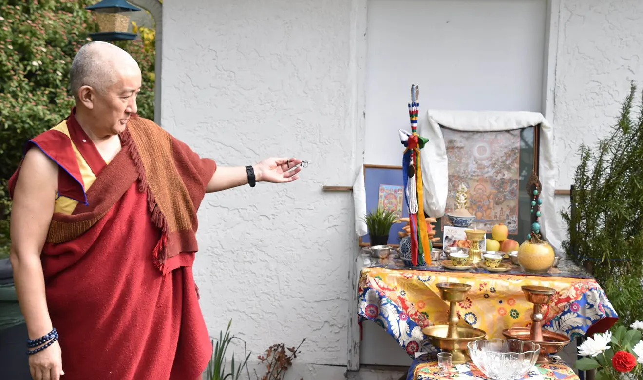 A Tibetan monk in traditional red robes stands by an ornate home altar. This sacred display is a central part of a Tibet cultural tour during Losar.
