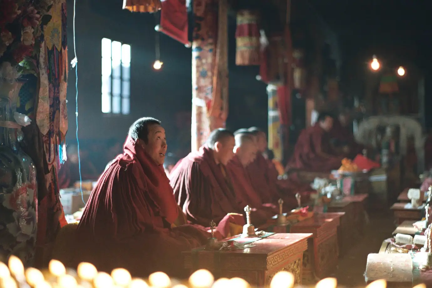 Tibetan Buddhist monks chant prayers inside a dimly lit monastery hall during a Tibet spiritual tour, surrounded by butter lamps, prayer texts, and colorful religious banners.