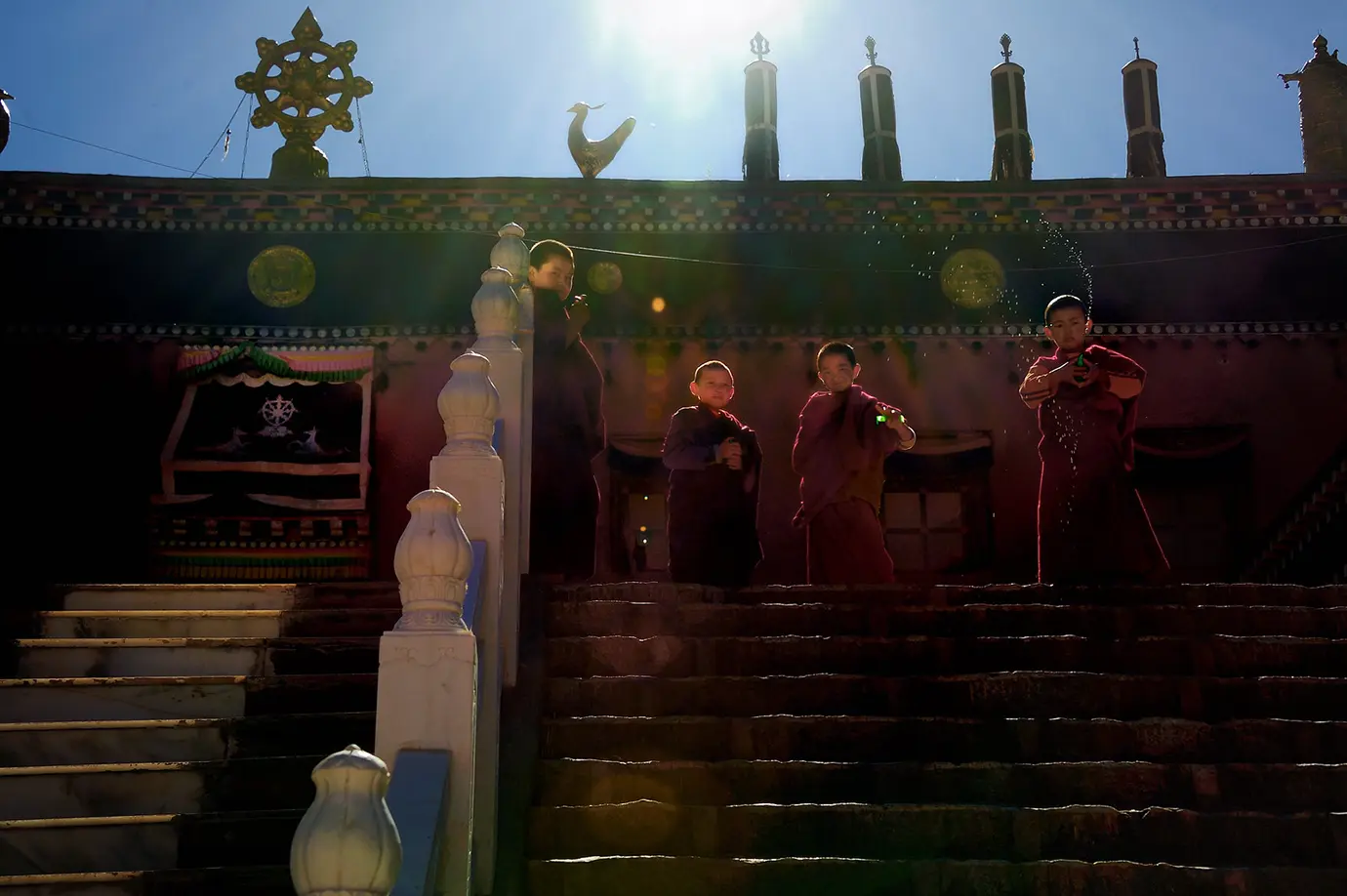 Young Tibetan monks stand on monastery steps in the Kham region with traditional temple symbols behind them, a cultural moment travelers may witness during a Tibet adventure tour.