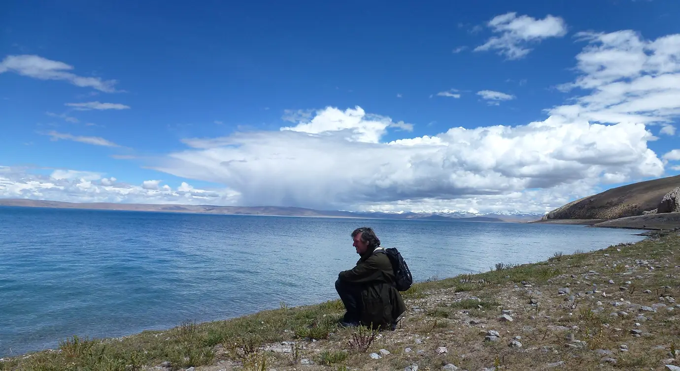 Traveler resting by the lakeshore during a Tibet Tour with Yamdrok Lake and Gyantse, with deep blue water, open grassland, and dramatic cloud-filled skies.