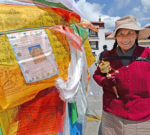 A smiling Tibetan senior woman holds a prayer wheel beside colorful prayer flags in a temple courtyard, showcasing a highlight of a Tibet tour for seniors.