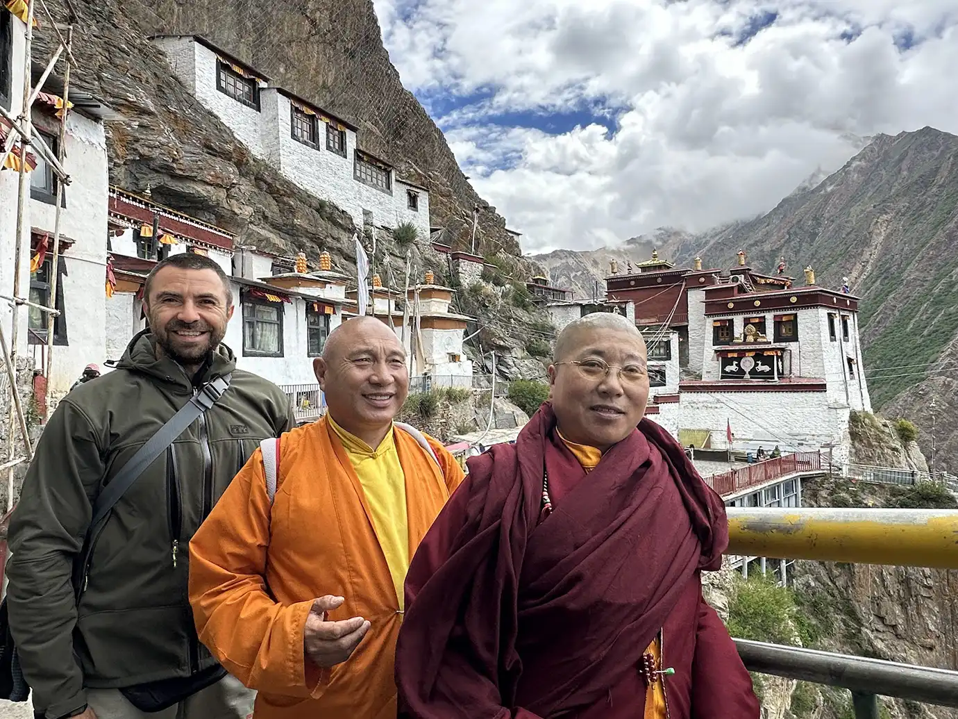 Travelers stand with Buddhist monks near a cliffside monastery during a Tibet Tour Safety-focused visit in the mountains, showing guided travel in a remote high-altitude setting.