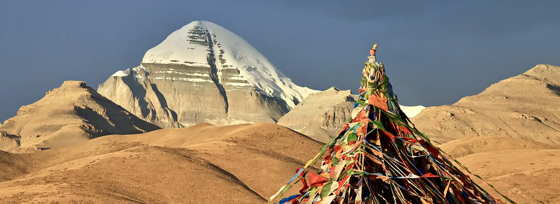 Vibrant prayer flags stand in a tall stack against the snow-capped peak of Mount Kailash. This iconic spiritual site is a major factor in overall Tibet tour cost.
