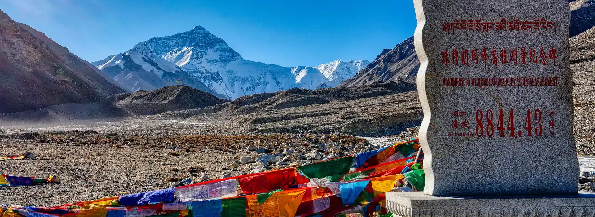 View of Mount Everest from the Tibetan side near Qomolangma Base Camp with prayer flags and a stone monument, a dramatic highlight of a Tibet adventure tour.