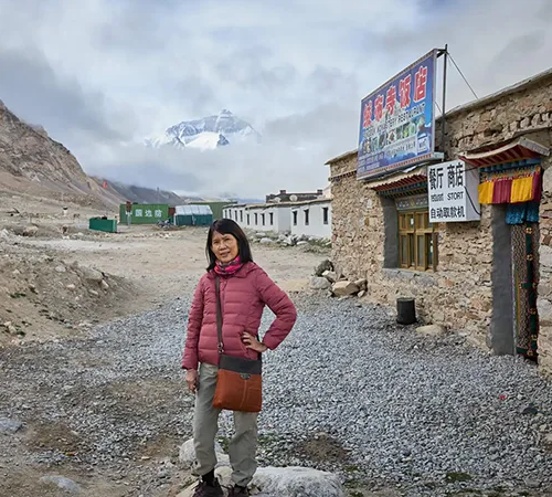 A woman in a pink puffer jacket stands on a gravel path near a stone building. Mount Everest appears behind her, illustrating a highlight of the Tibet tour cost.