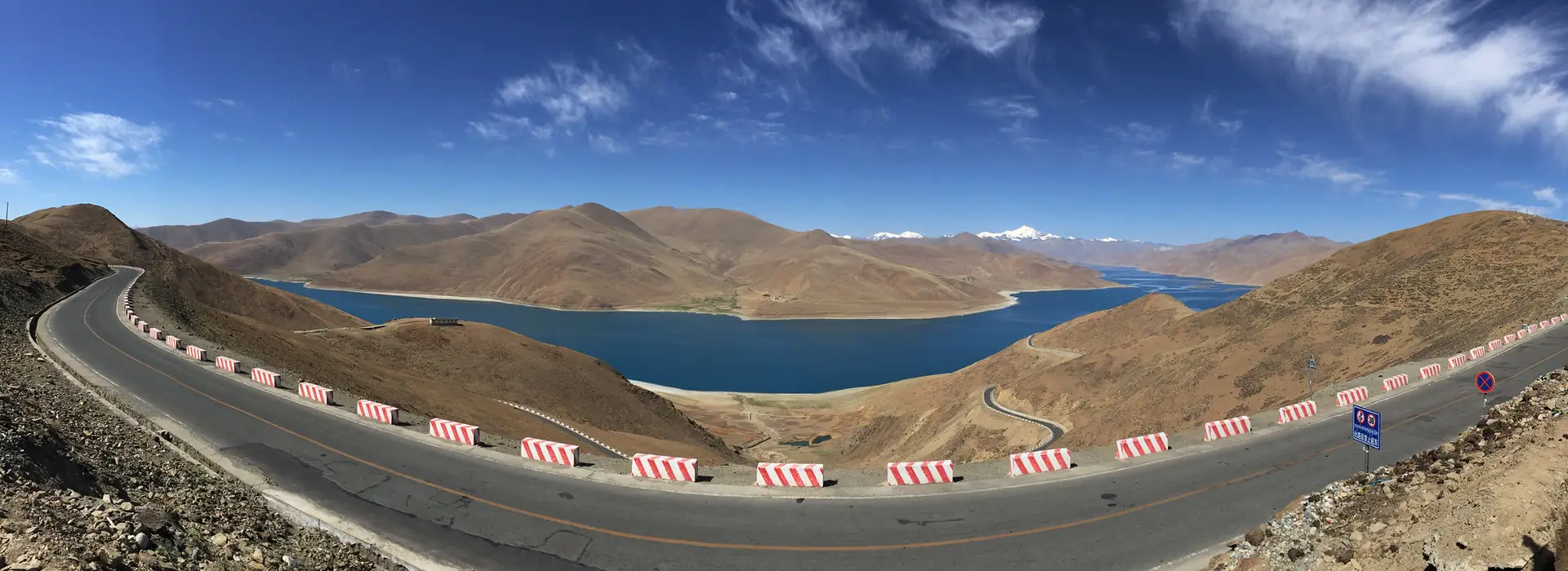 A high mountain road curves above a deep blue lake in Tibet, showing the scenic overland routes travelers often research when learning how to get to Tibet.