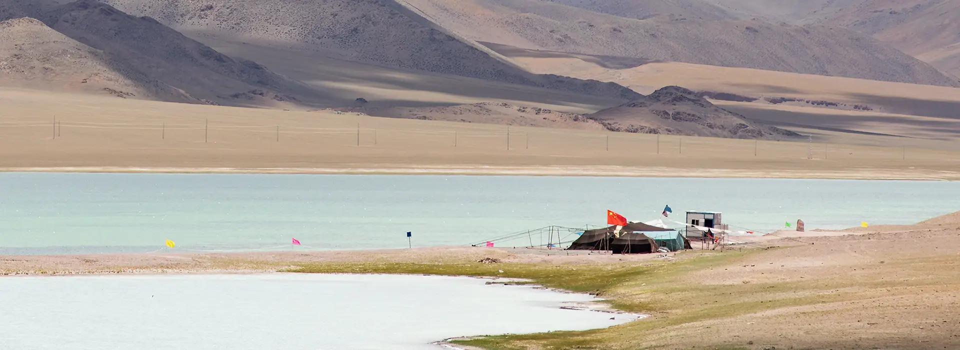 A scenic turquoise lake sits before vast rolling mountains. A small campsite with flags marks the shore, a serene stop on a Tibet tour for seniors.