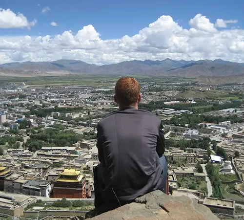 A solo traveler sits on a high viewpoint above Lhasa, looking across the city and distant mountains while planning a safe solo Tibet travel itinerary.