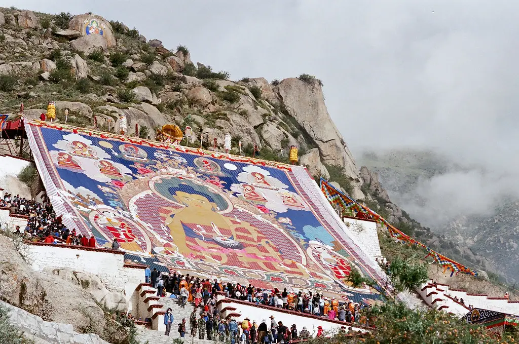 A massive thangka is displayed on a hillside while a large crowd of pilgrims gathers below, a highlight of a Tibet Buddhist Pilgrimage Tour.