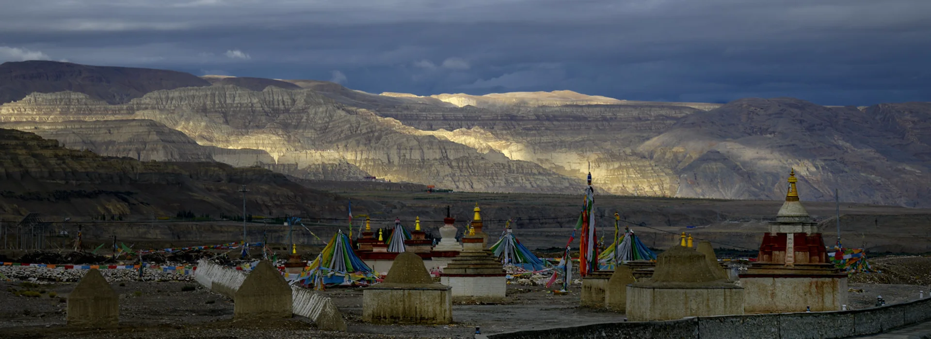 Stupas and prayer flags stand beneath dark clouds and sunlit mountains during a Tibet Tour Safety route through a remote high-altitude valley.