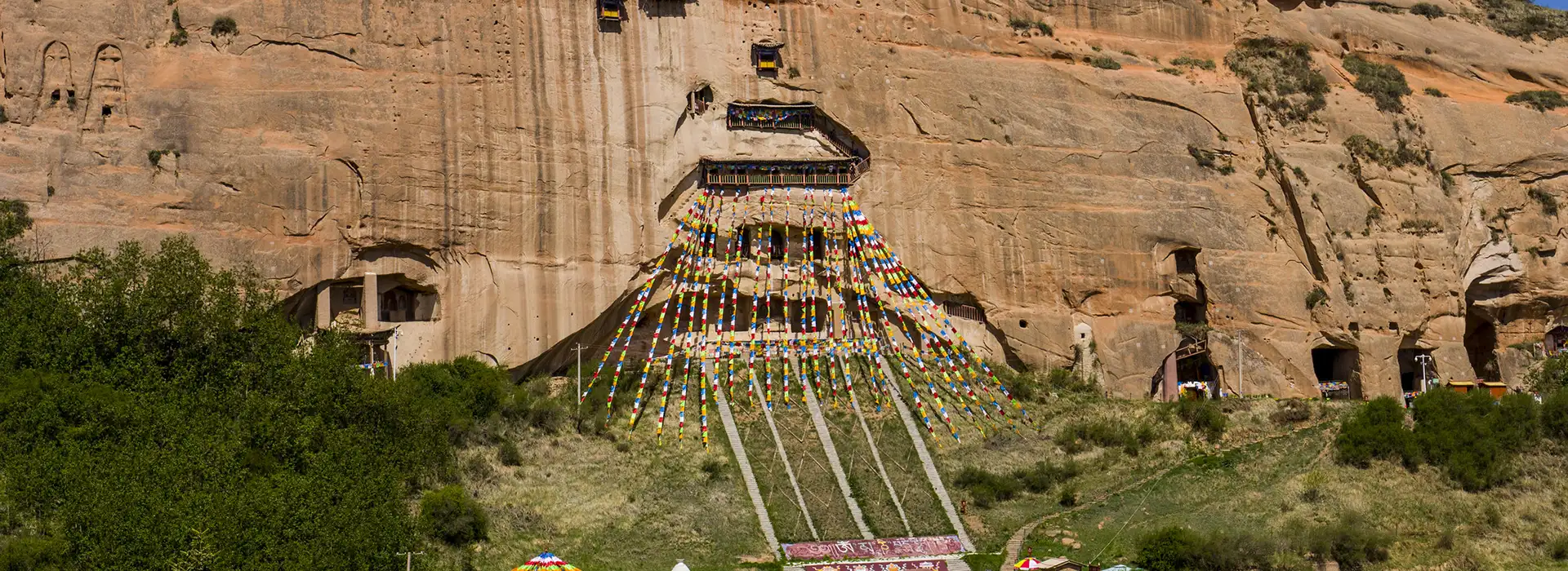 Colorful prayer flags cascade down a cliffside monastery carved into rock during a Tibet spiritual tour, where travelers visit sacred caves and ancient Buddhist meditation sites.
