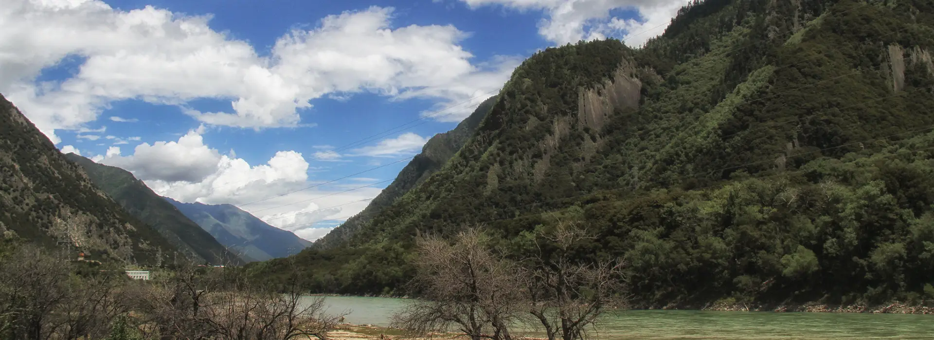 River valley landscape on a Tibet Tour with Yamdrok Lake and Gyantse, featuring forested mountain slopes, calm water, and a wide blue sky with large white clouds.