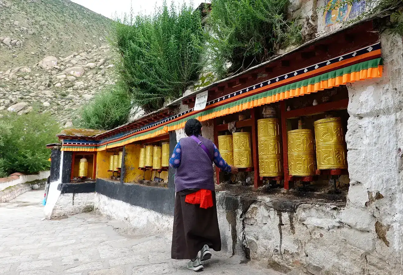 A woman in traditional dress spins a row of large golden prayer wheels at a monastery. This spiritual experience is a highlight of a budget Tibet tour.