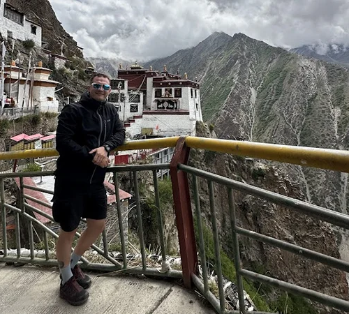 A traveler in hiking gear stands on a mountain balcony overlooking a historic cliffside monastery during a Tibet tour with Yamdrok Lake and Gyantse.
