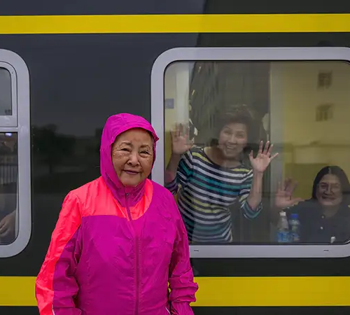 A smiling traveler in a bright pink jacket stands beside a train window while passengers wave from inside during a Tibet tour by train across the high plateau.