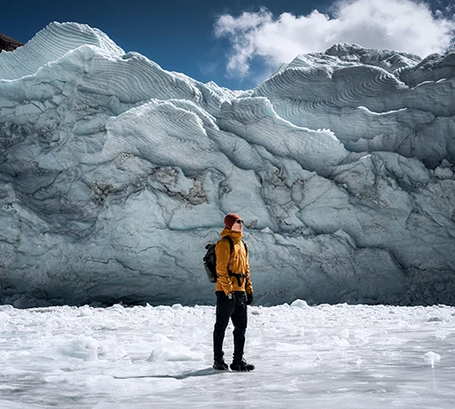 A lone hiker in a yellow jacket stands before a massive, towering ice glacier with intricate textures, a highlight of adventure-focused Tibet trekking tours.