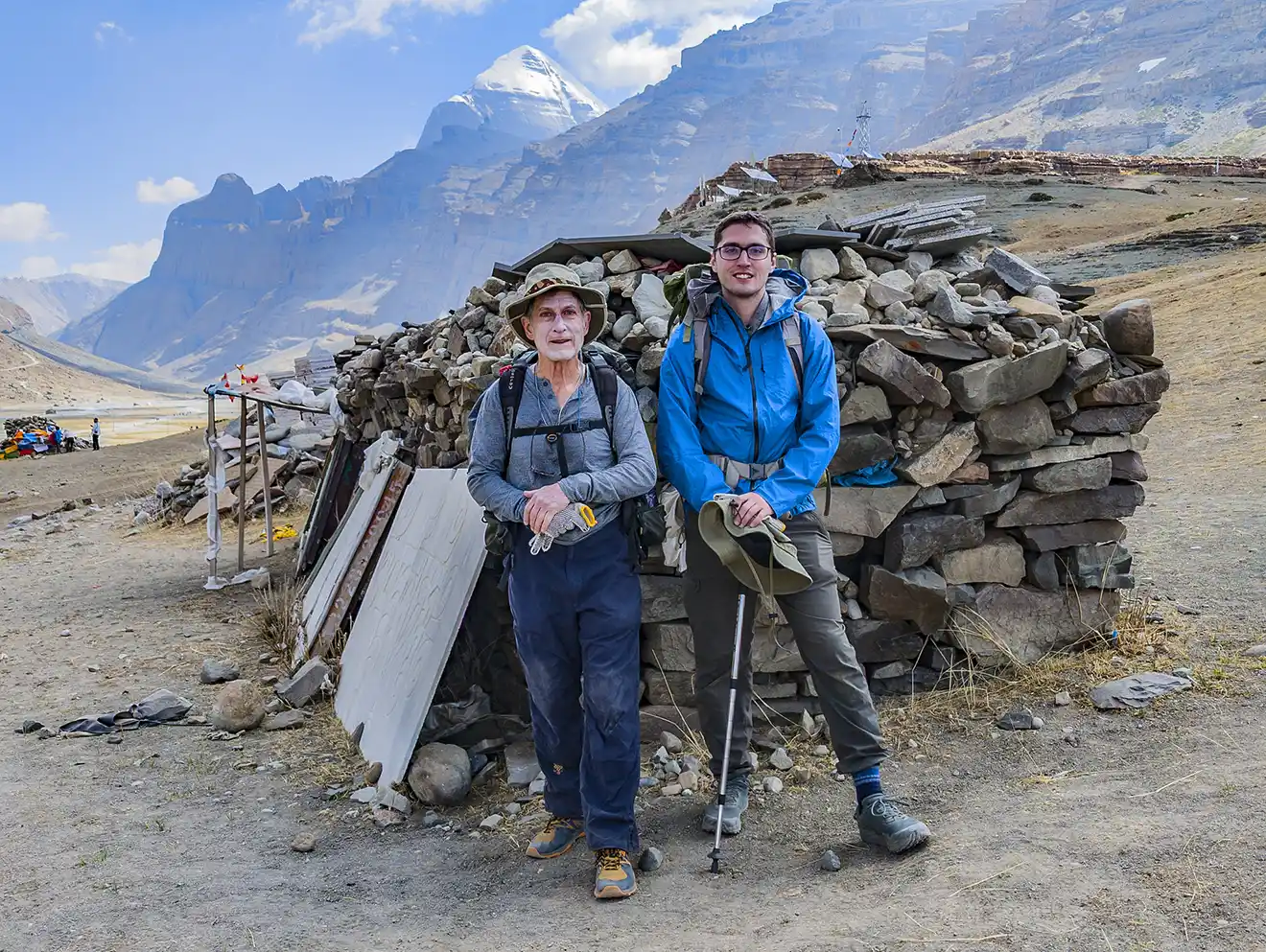Two hikers stand before a stone structure with Mount Kailash in the distance. This rugged landscape is a highlight of a Tibet tour for seniors.