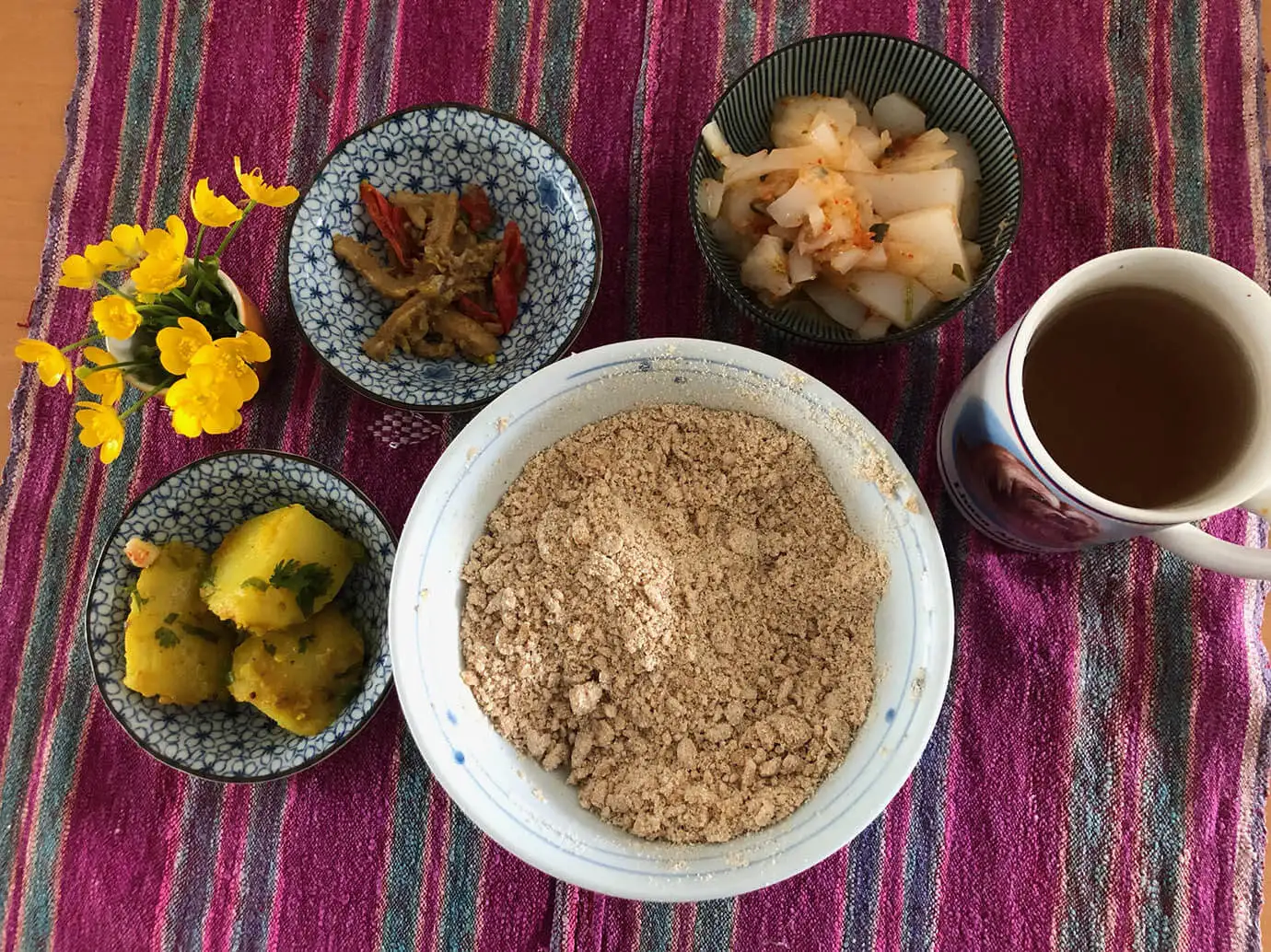 A bowl of traditional Tsampa sits alongside small dishes of potatoes and tea on a colorful textile, essential sustenance during a Tibet Buddhist Pilgrimage Tour.