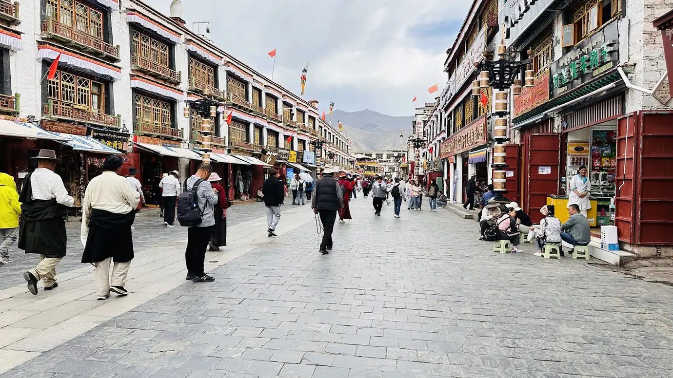 Travelers and locals walk along a busy street in Lhasa during a Tibet Tour Safety-focused city visit near shops, prayer routes, and traditional Tibetan buildings.