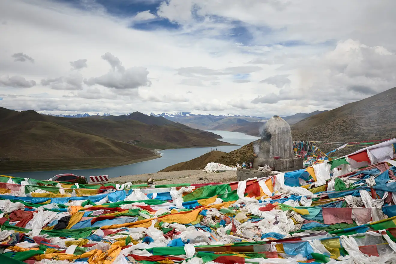 Colorful prayer flags fill the foreground at a high mountain pass. A distant blue lake and snow-capped peaks are viewed on a Tibet tour for seniors.