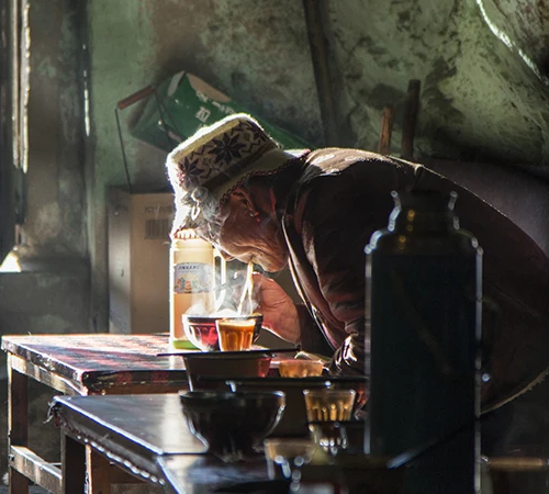 An elder in a traditional hat eats a steaming bowl of authentic Tibetan food inside a rustic local eatery filled with natural morning light.