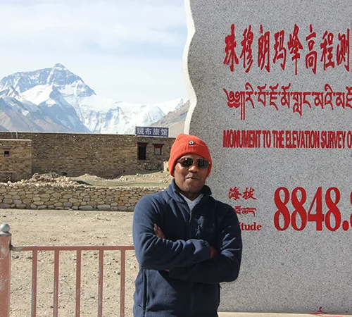 A smiling traveler poses beside the 8848.86m elevation marker monument, with Mount Everest visible behind him, during a Lhasa to Everest Base Camp Tour.