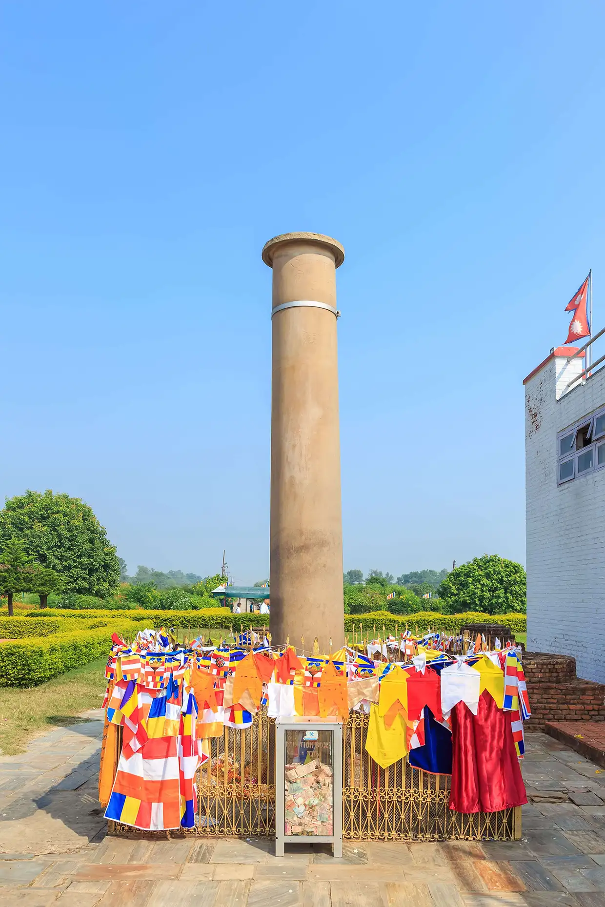 The ancient sandstone Ashoka Pillar stands tall under a clear blue sky, surrounded by colorful prayer flags. This landmark is essential for any Lumbini Travel Guide.