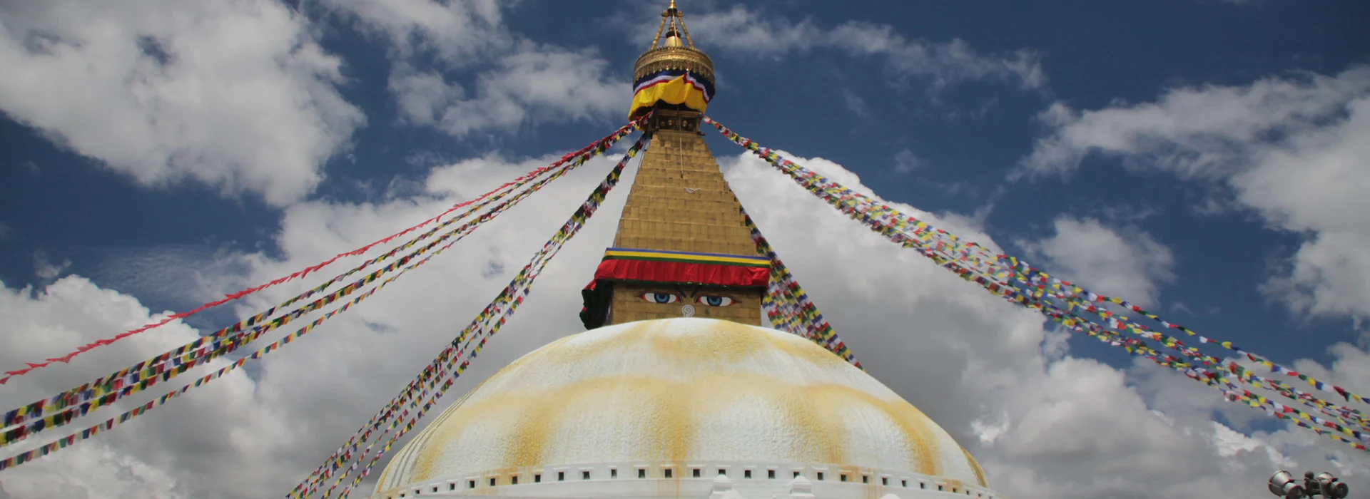 The golden spire of Boudhanath Stupa rises against a cloudy sky. Colorful prayer flags stretch across the dome as visitors plan a trip to Nepal.