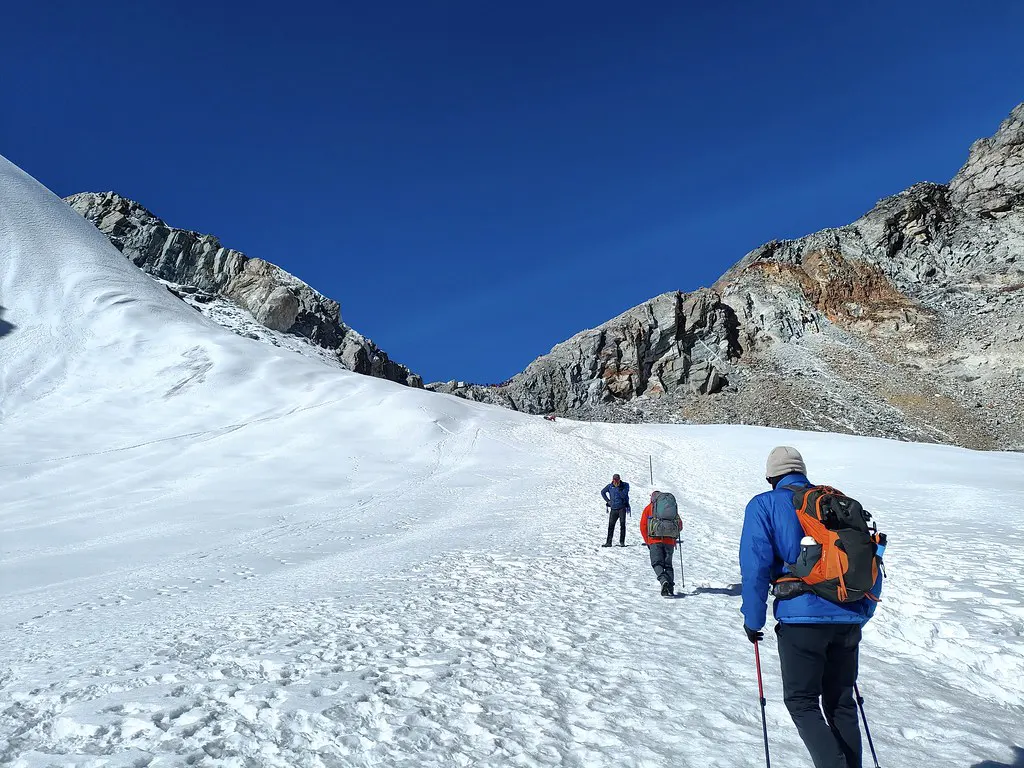 Three trekkers in insulated winter gear and backpacks ascend a steep, snow-covered glacier toward a rocky mountain pass under a clear blue sky during an Everest Three Passes Trek.