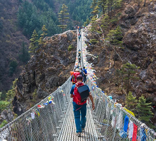 A group of trekkers crosses a long suspension bridge draped with prayer flags over a deep forested gorge, following an expert Gokyo Lakes Trek Guide.